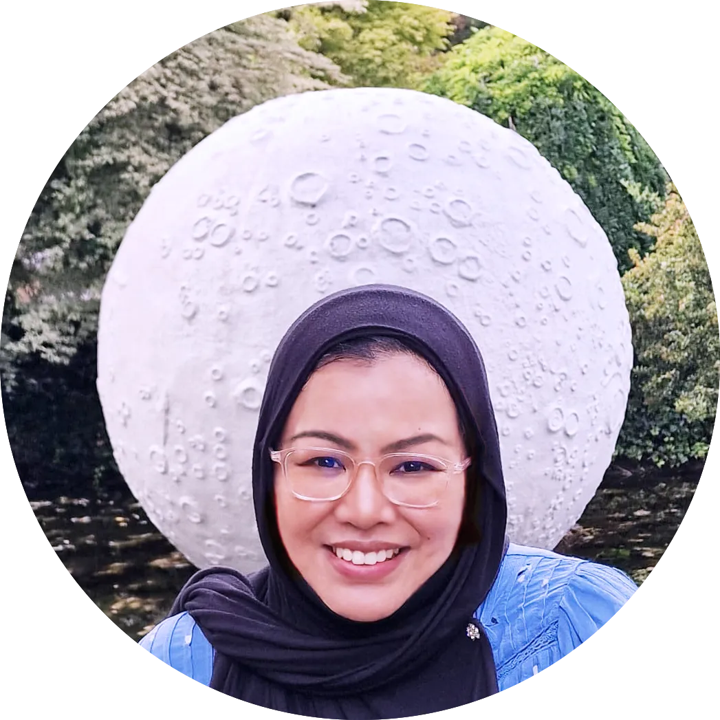 Author picture. Behind her head, a moon sculpture is visible in a backdrop of trees. She looks happy for some reason!