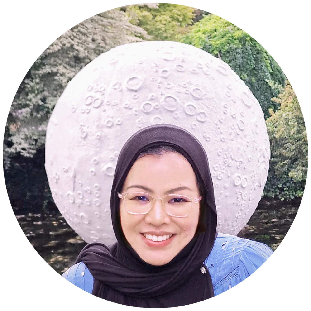 Author picture. Behind her head, a moon sculpture is visible in a backdrop of trees. She looks happy for some reason!